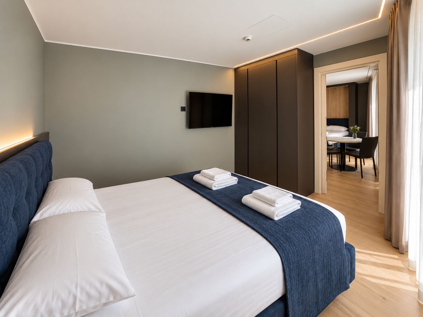 Habitación de hotel moderna con una cama grande, un televisor de pantalla plana montado en la pared y un área de comedor separada visible a través de una puerta abierta.
