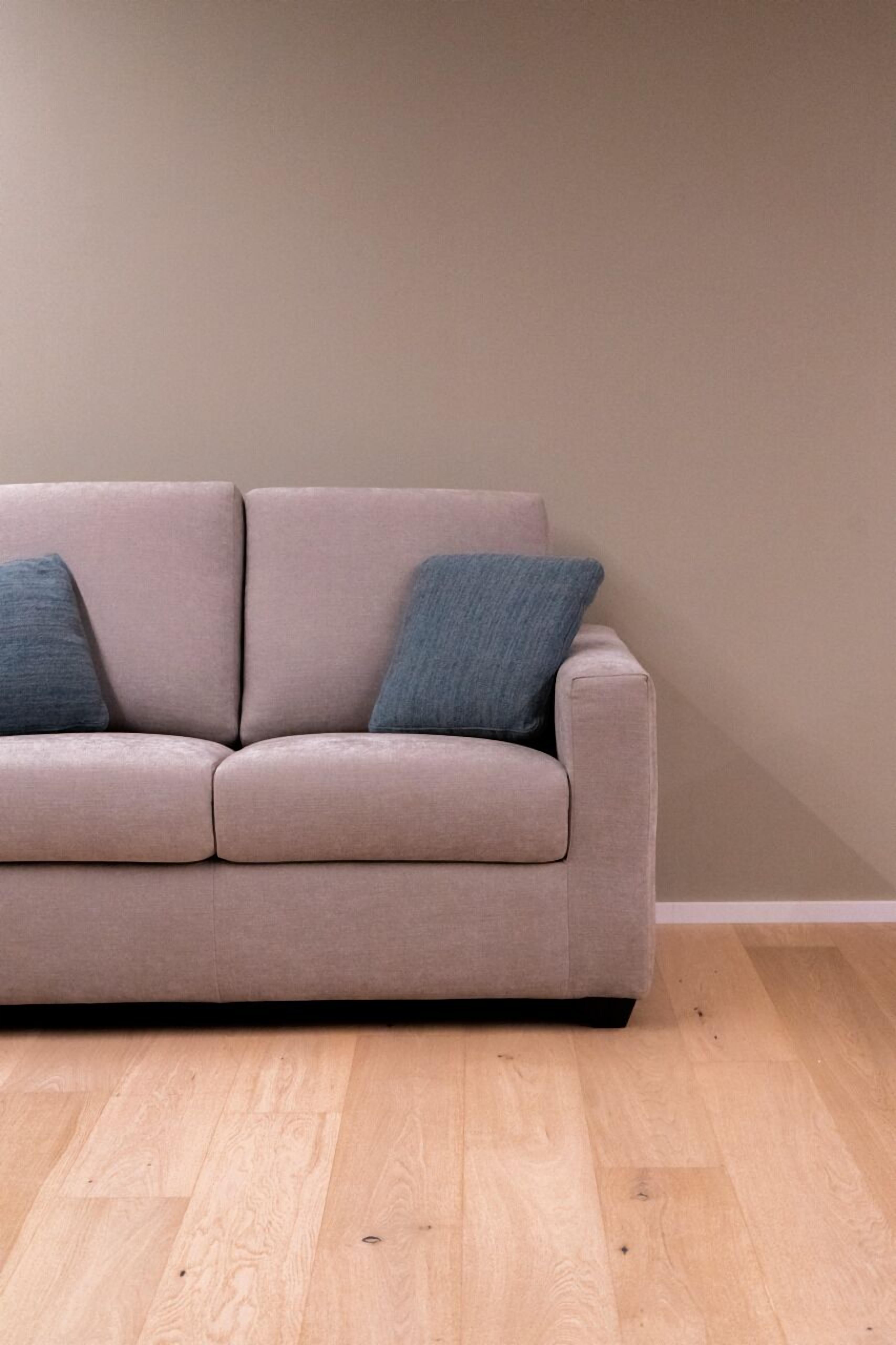 Un sofá beige con dos cojines oscuros frente a una pared lisa y suelo de madera.