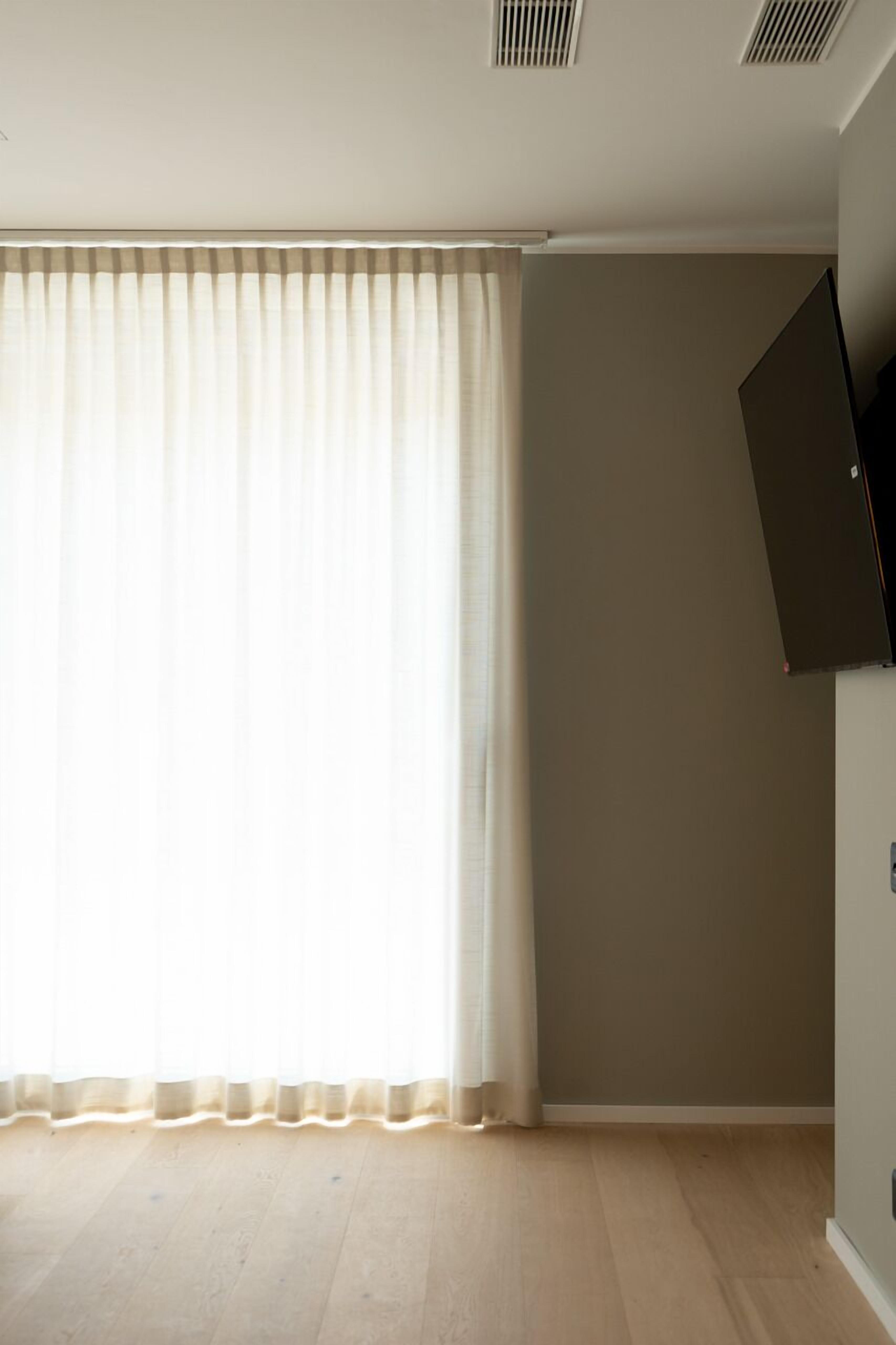 A room with a large window covered by sheer curtains, a flat-screen TV mounted on the wall, and a wooden floor.