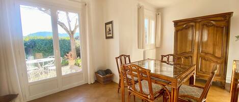 Un coin repas avec une table en verre et des chaises en osier, de grandes fenêtres avec des rideaux et une vue sur la verdure environnante.