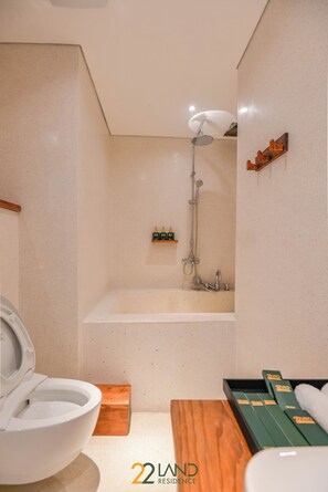 A bathroom with a toilet, a sink, a bathtub, and a shower. There are toiletries on a shelf and a towel on a wooden bench. - 22land Residence Suite (Hanoi)