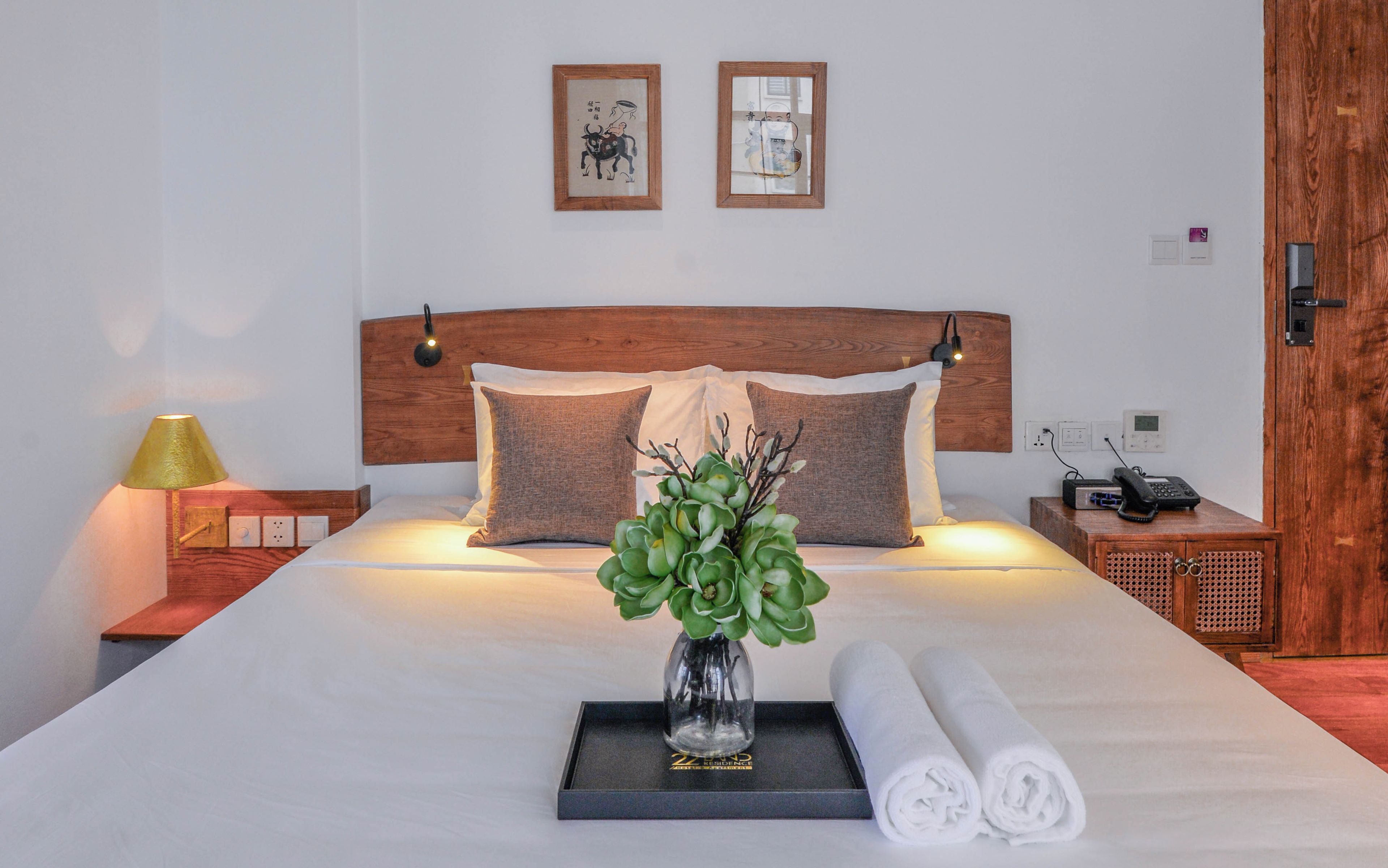 A neatly made bed with a vase of green plants, rolled towels, and two framed artworks on the wall.