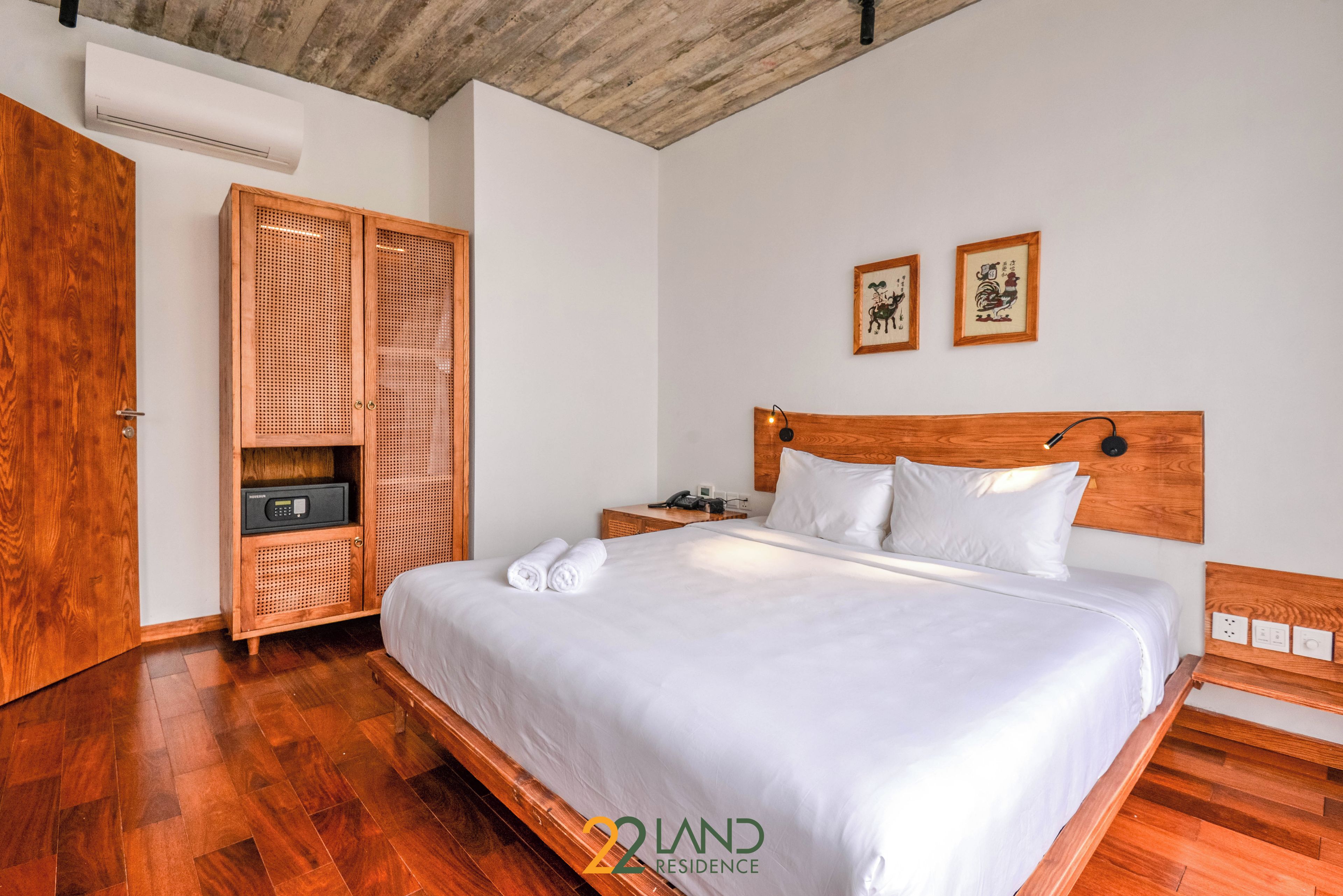 A neatly made bed with white linens, a wooden headboard, and a bedside table with a lamp. A wooden wardrobe with a woven door is beside the bed.