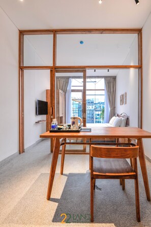 A modern hotel room with a wooden dining table, a single chair, a bed with white linens, a TV mounted on the wall, and a large window offering a city view. - 22land Residence Suite (Hanoi)
