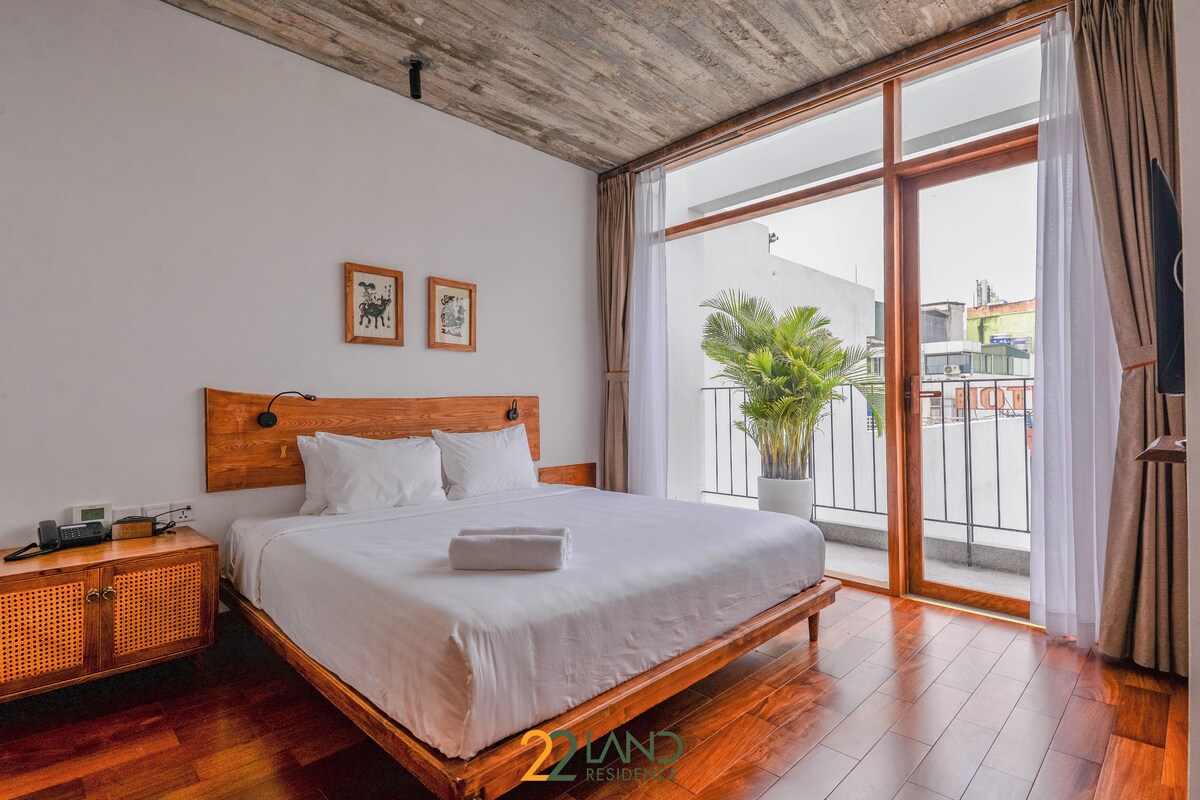 A bedroom with a wooden bed, white bedding, a nightstand, a large window with a view of the outside, and a ceiling with exposed beams.