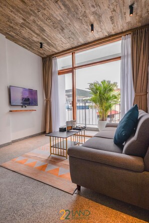 A modern living room with a sofa, a flat-screen TV mounted on the wall, a potted plant, and a view through large windows. - 22land Residence Suite (Hanoi)