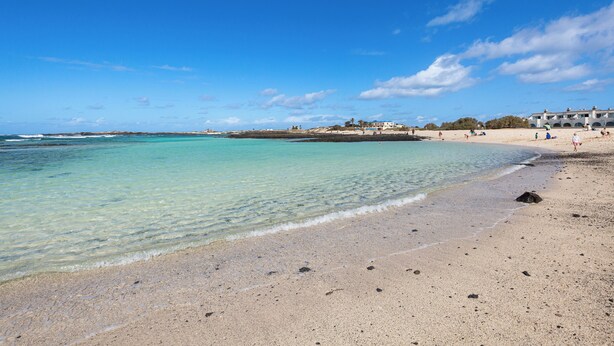 Une plage avec une eau turquoise et limpide, un rivage sablonneux et, au loin, un bâtiment à plusieurs étages.