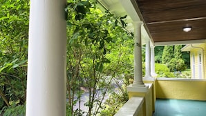 Een overdekte veranda met witte zuilen en een gele muur, omgeven door weelderig groen.
