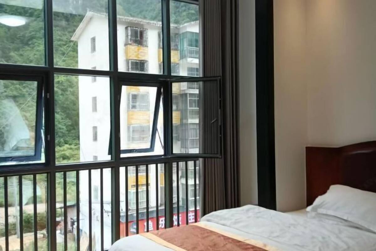 A hotel room with a large window, a bed with white bedding, and a view of buildings and greenery outside.
