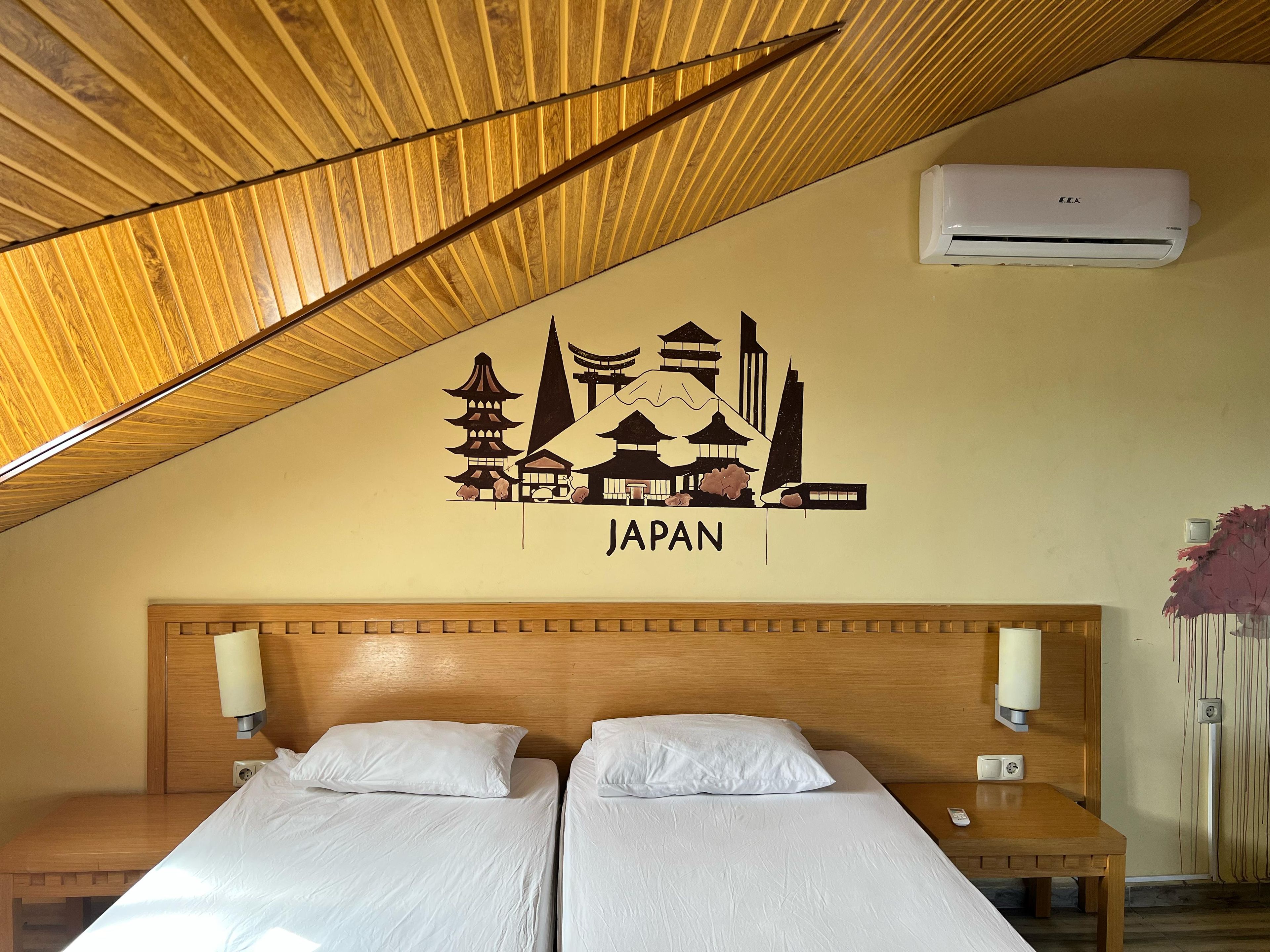 A hotel room with a wooden sloped ceiling, two beds with white bedding, a wall-mounted air conditioner, and a Japan-themed wall decal.