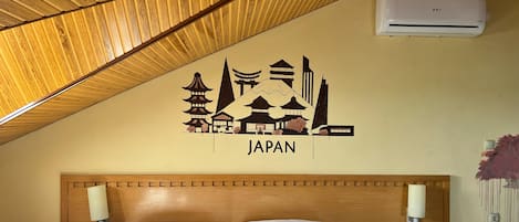 A hotel room with a wooden sloped ceiling, two beds with white bedding, a wall-mounted air conditioner, and a Japan-themed wall decal.