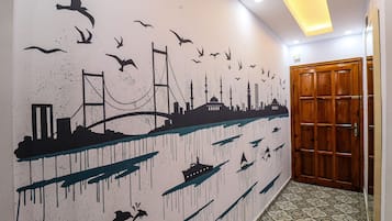 Un pasillo con un mural que representa un horizonte urbano con un puente, edificios y pájaros volando. A la derecha, se observa una puerta de madera.