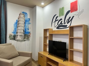 A living room with a beige sofa, a flat-screen TV on a wooden stand, and a wall mural of the Leaning Tower of Pisa. - Esma Apart Hotel (Alanya)