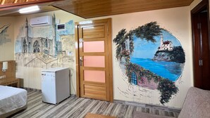 A room with a wooden ceiling, a door with pink panels, a refrigerator, a bed, and a wall mural of a coastal scene. - Esma Apart Hotel (Alanya)
