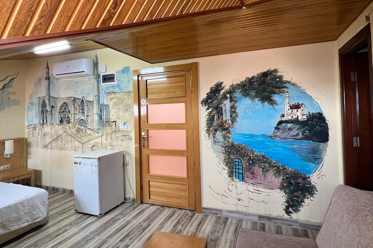 Une pièce avec un plafond en bois, une porte à panneaux roses, un réfrigérateur, un lit et une fresque murale représentant un paysage côtier.