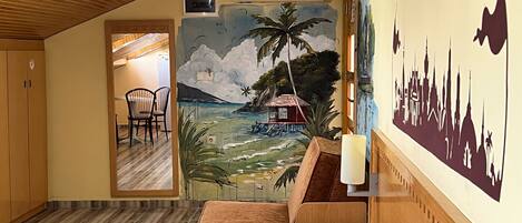 A hotel room with a bed, wooden furniture, a mural of a tropical beach scene, and a city skyline silhouette on the wall.