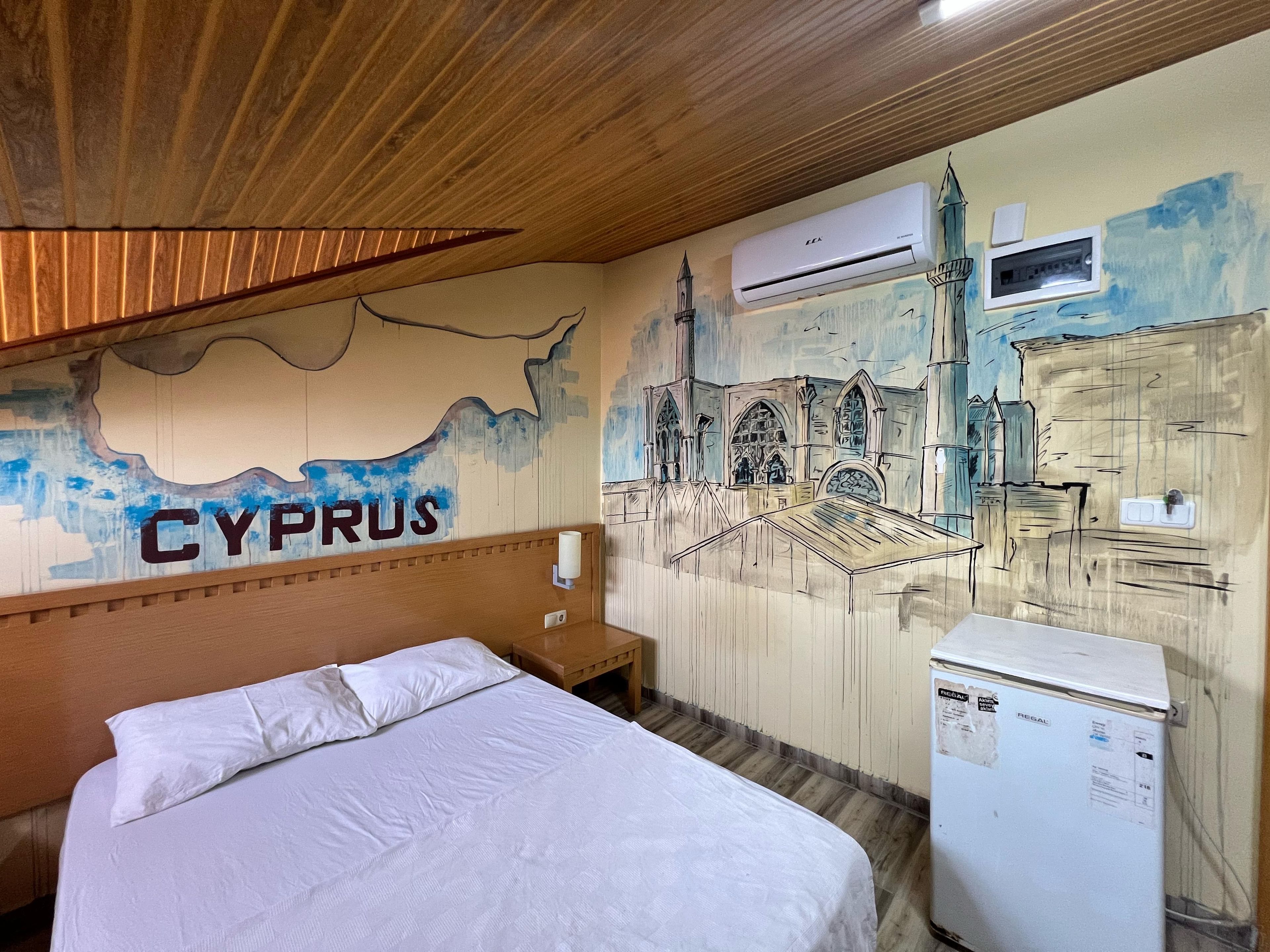 A hotel room with a bed, a wooden headboard, a wall mural of a cityscape, and a refrigerator.