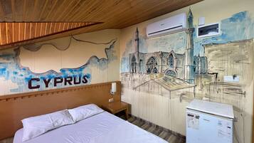 A hotel room with a bed, a wooden headboard, a wall mural of a cityscape, and a refrigerator.