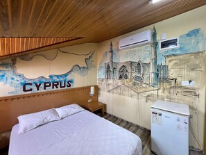 A hotel room with a bed, a wooden headboard, a wall mural of a cityscape, and a refrigerator.