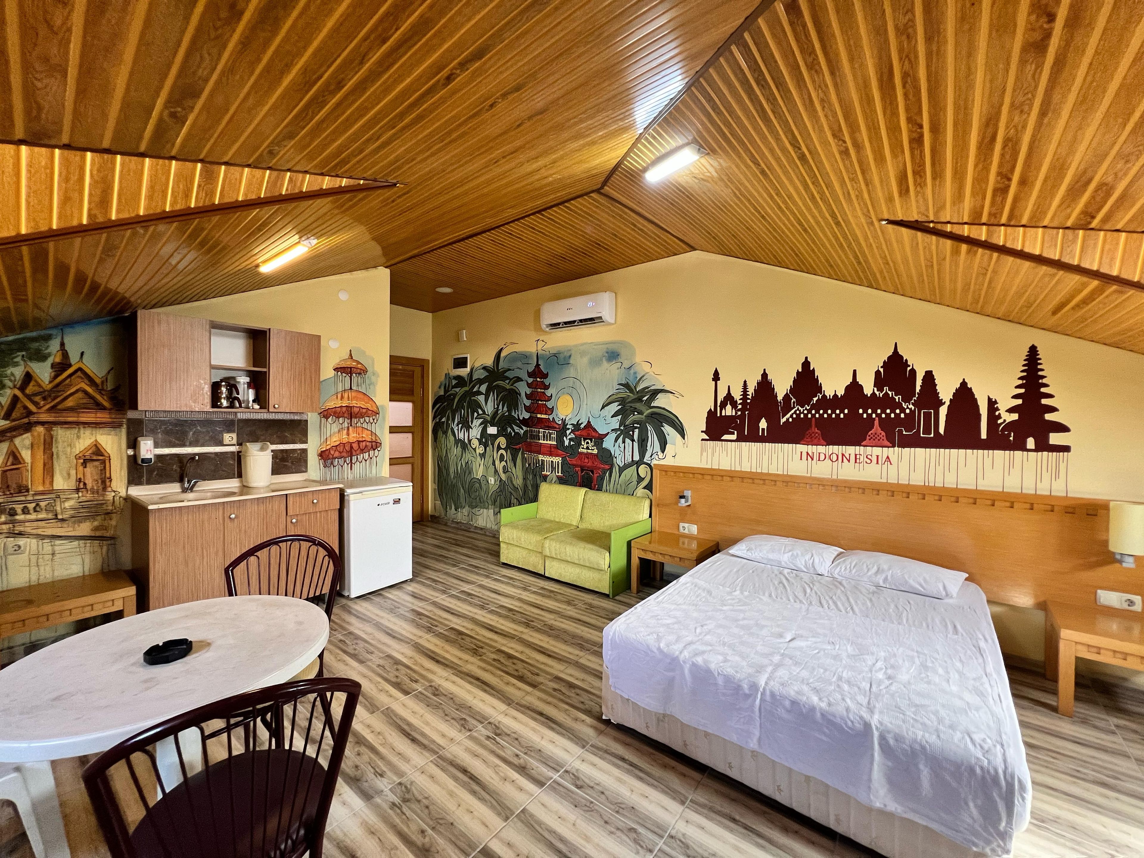 A hotel room with a bed, a desk with a chair, a small kitchenette, and a mural of Indonesian landmarks.