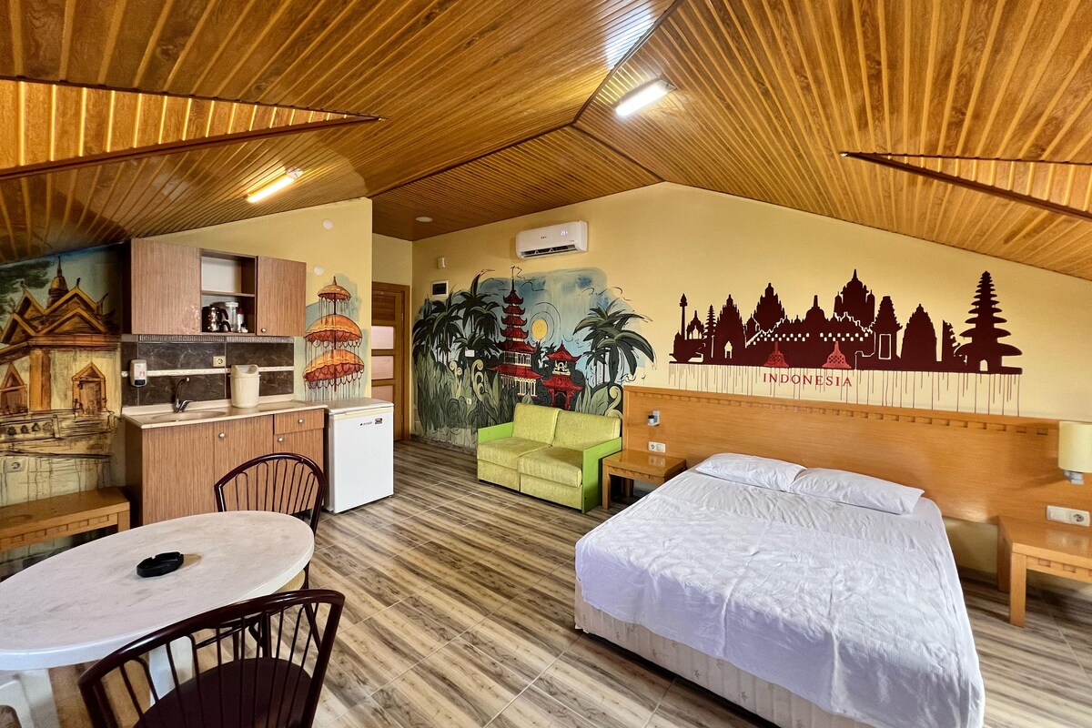 Une chambre d’hôtel comprenant un lit, un bureau avec une chaise, une petite kitchenette et une fresque murale représentant des monuments indonésiens.