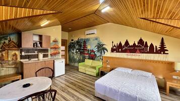 A hotel room with a bed, a desk with a chair, a small kitchenette, and a mural of Indonesian landmarks.
