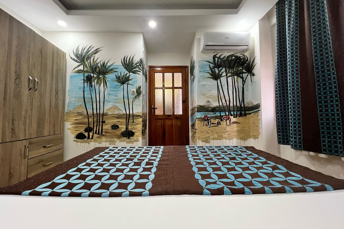 Une pièce avec une armoire en bois, un tapis à motifs et des peintures murales représentant des palmiers et des pyramides.
