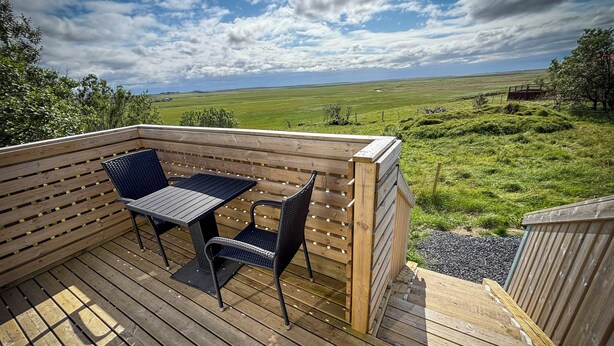 Una terraza de madera con una mesa y dos sillas, con vista a un campo verde y un cielo nublado.