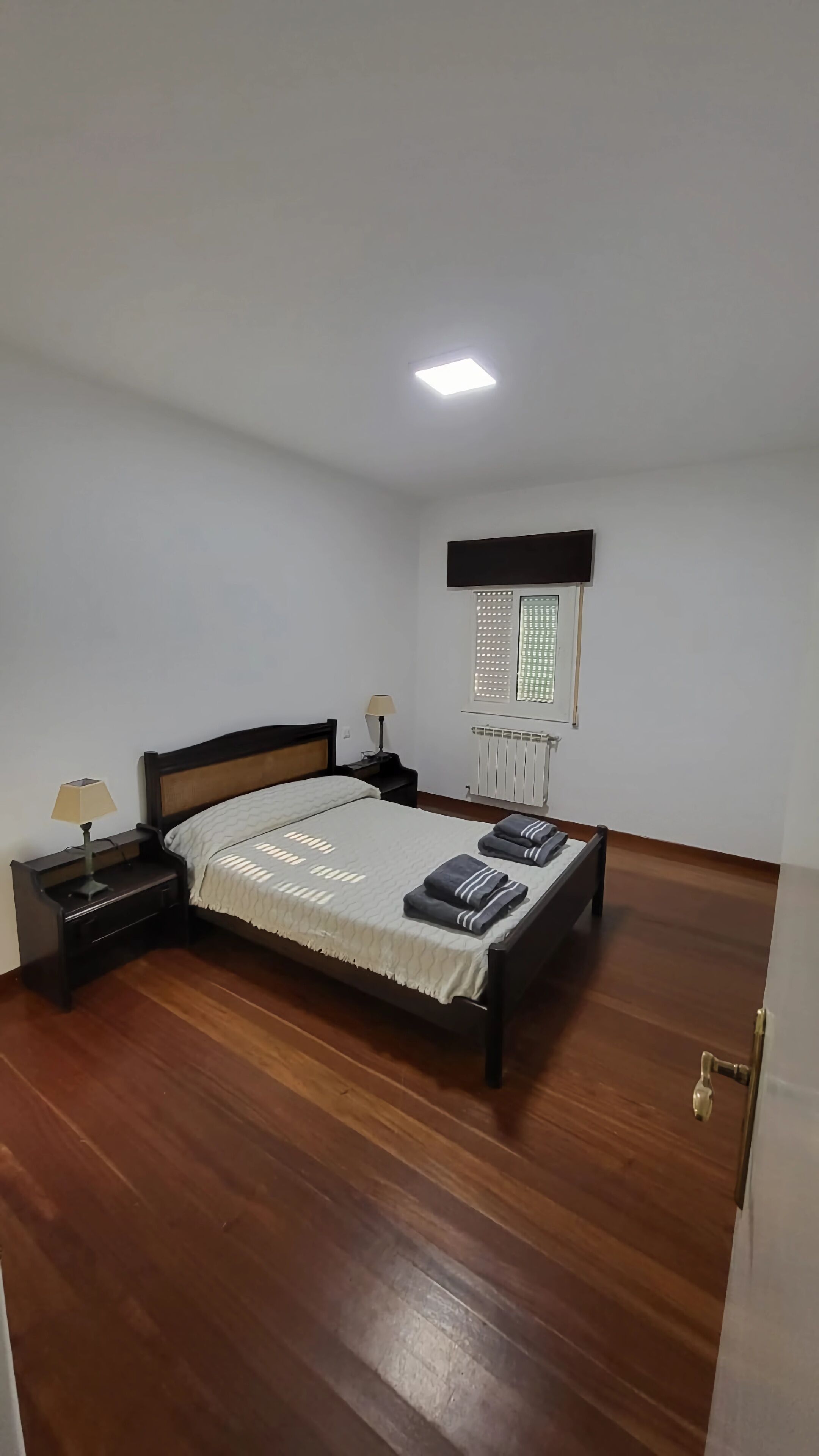 Een slaapkamer met een houten vloer, een bed met hoofdeinde, een nachtkastje met een lamp, een raam met jaloezieën en een radiator.