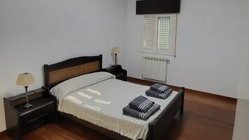 Een slaapkamer met een houten vloer, een bed met hoofdeinde, een nachtkastje met een lamp, een raam met jaloezieën en een radiator.