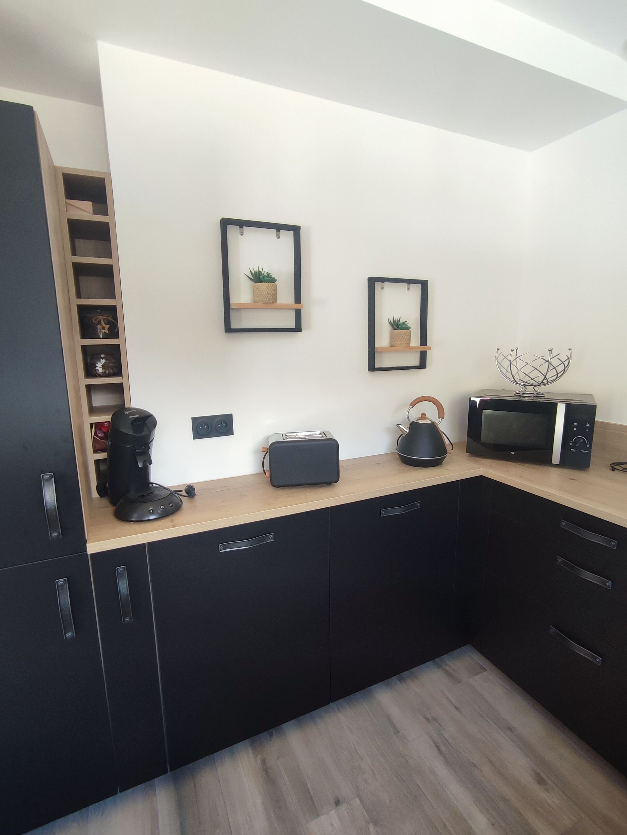 Una cocina moderna con armarios negros, una encimera de madera, cafetera, tostadora, hervidor y microondas. Hay dos cuadros en la pared.