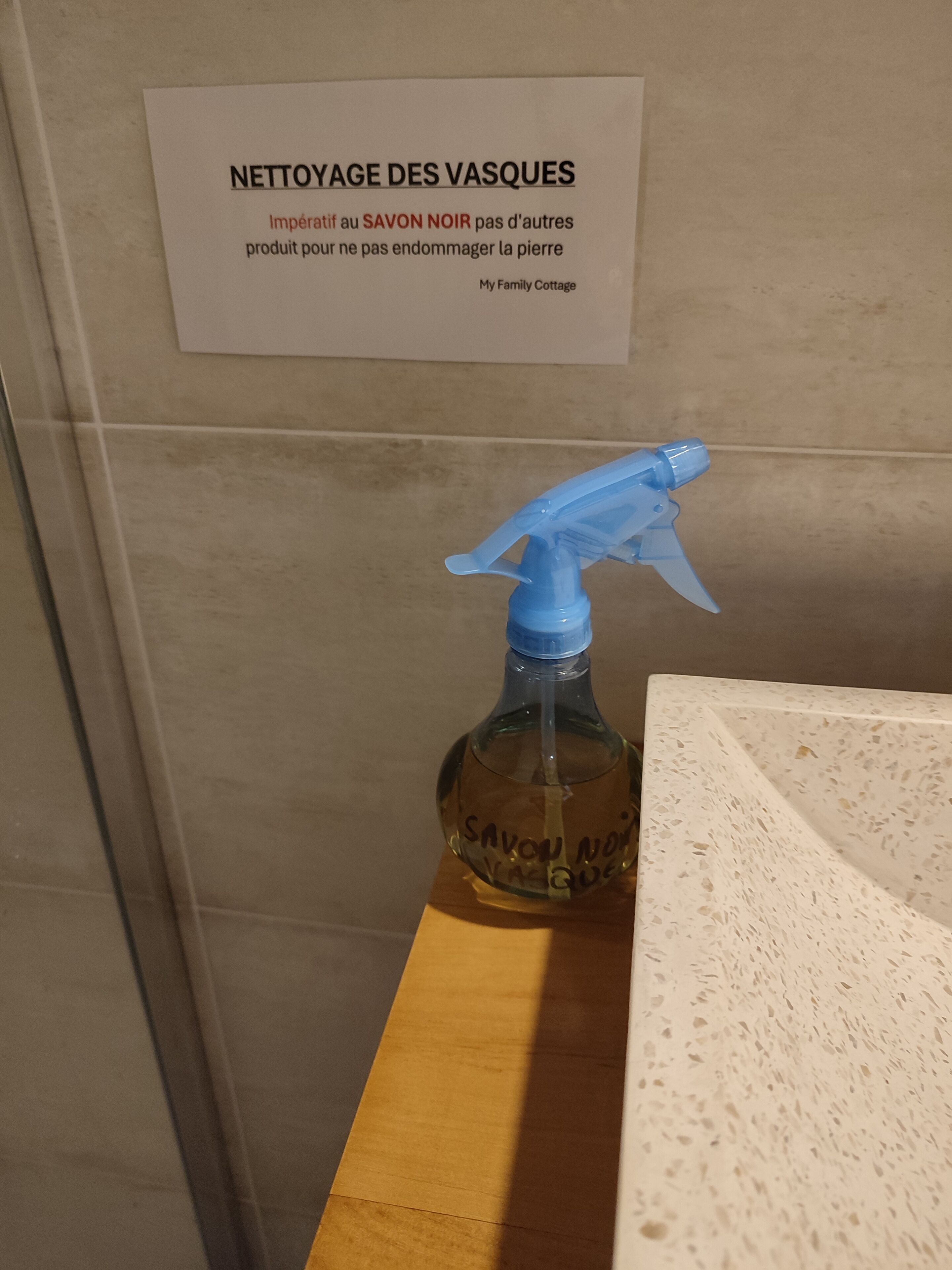 A bathroom with a sign above a sink that reads "NETTOYAGE DES VASQUES" and a bottle labeled "SAVON NOIR" with a spray nozzle.