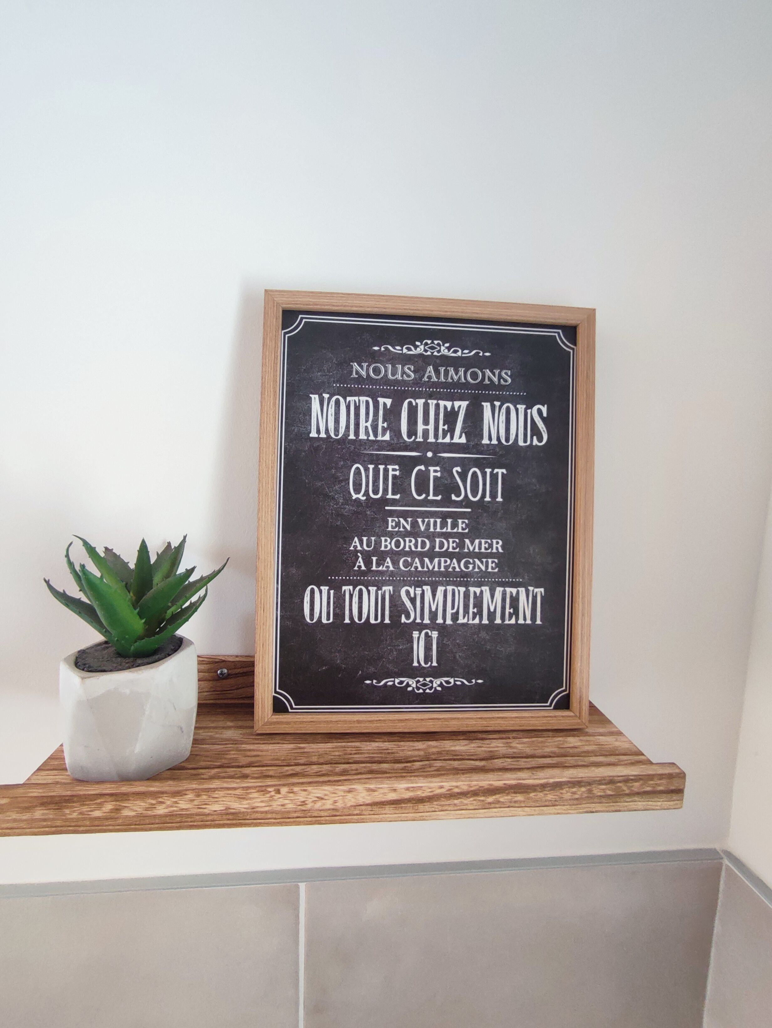 Une plaque encadrée portant un message en français, placée sur une étagère en bois, à côté d’une plante en pot.