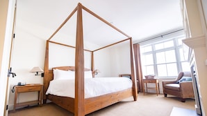 Una cama con dosel en una habitación de hotel, con cabecera de madera, una mesita de noche con lámpara, una silla y una ventana con cortinas.