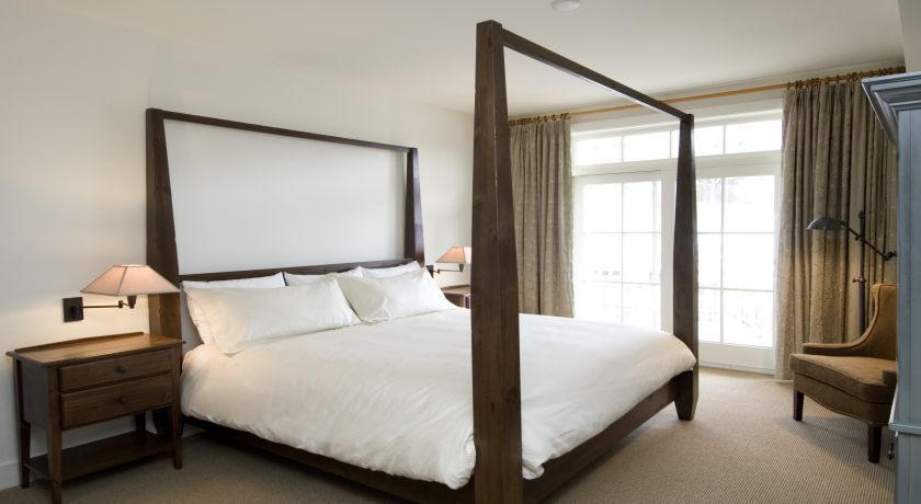 A four-poster bed with white linens, wooden nightstands with lamps, and a view of the outdoors through large windows.
