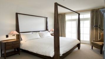 A four-poster bed with white linens, wooden nightstands with lamps, and a view of the outdoors through large windows.