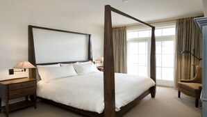Una cama con dosel adornada con sábanas blancas, mesitas de noche de madera con lámparas, y vistas al exterior a través de amplios ventanales.