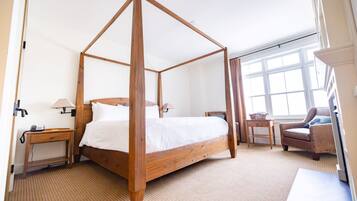 A four-poster bed in a hotel room with a wooden headboard, a bedside table with a lamp, a chair, and a window with curtains.
