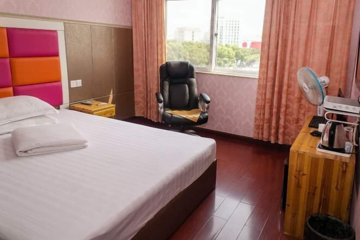 A hotel room with a bed, a chair, a desk with a fan, and a window with curtains.