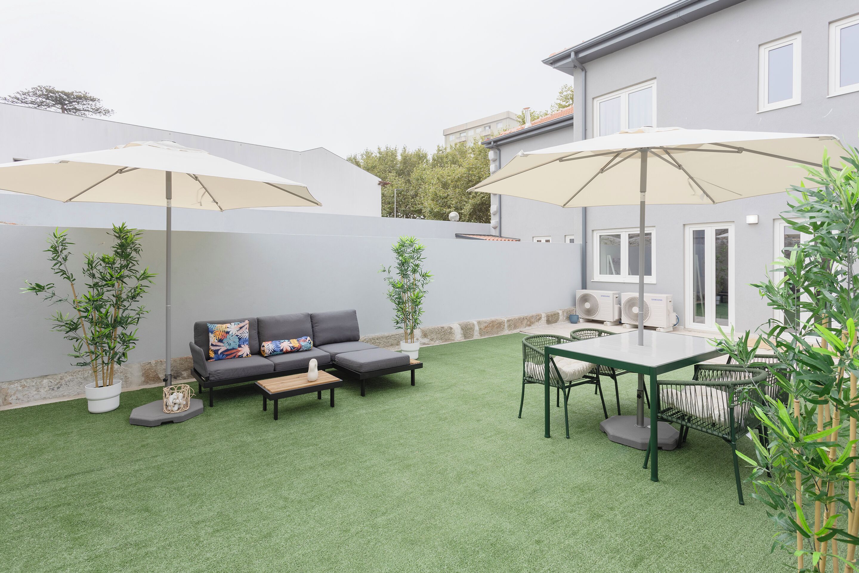 A patio with artificial grass, a sofa, two umbrellas, a table, chairs, and potted plants.