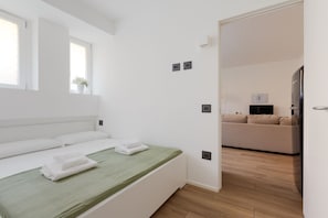 Una habitación de hotel moderna con una cama, un edredón verde, una ventana con una planta, un sofá en la habitación contigua y un televisor de pantalla plana.