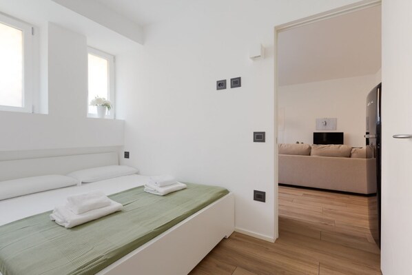 Una habitación de hotel moderna con una cama, un edredón verde, una ventana con una planta, un sofá en la habitación contigua y un televisor de pantalla plana.