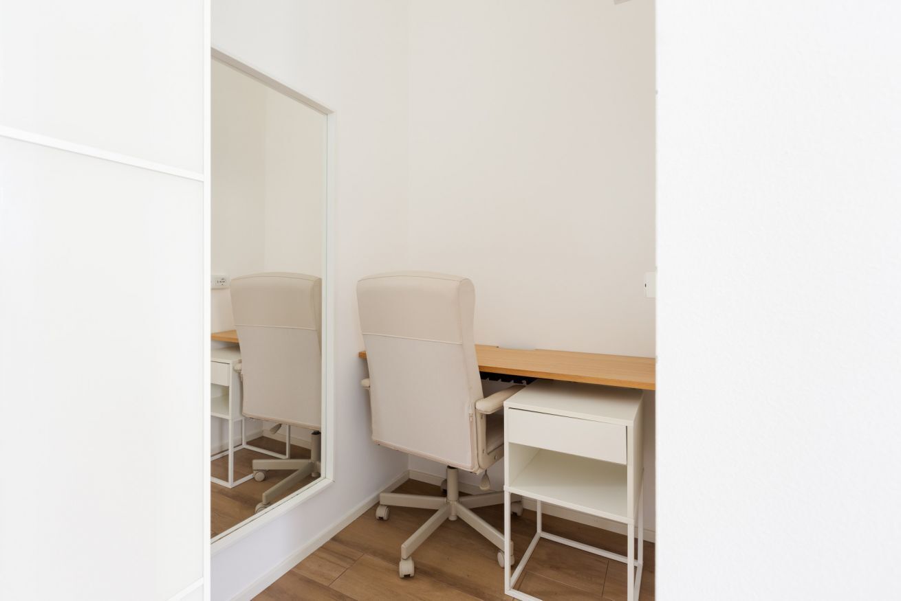A small, white room with a desk and chair, a mirror, and a wooden floor.