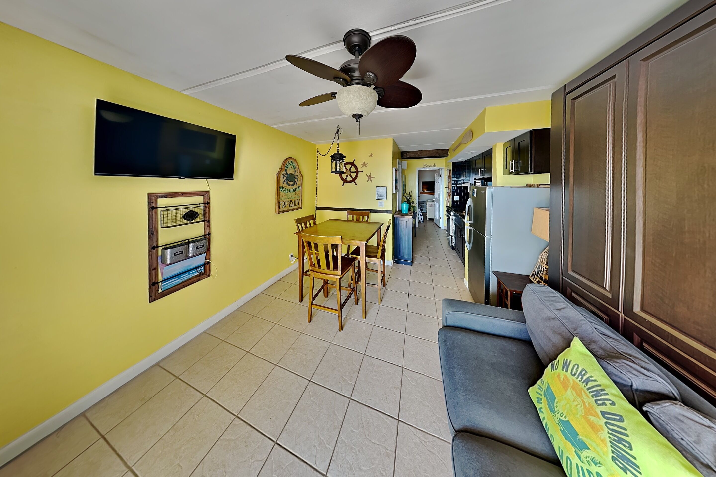 A living room with a sofa, a dining table with chairs, a ceiling fan, and a TV mounted on the wall.
