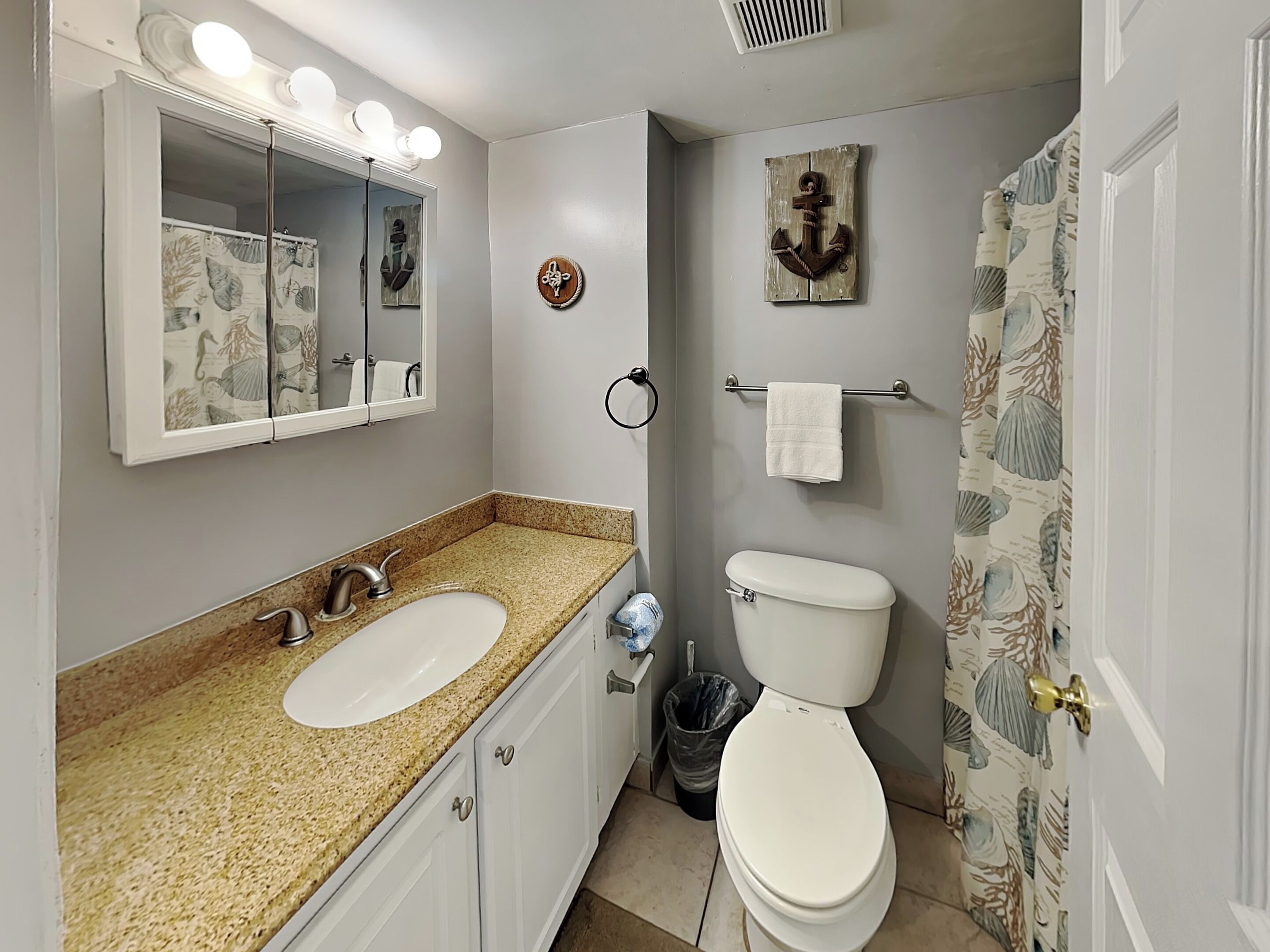 A bathroom with a granite countertop sink, a mirror with lights, a toilet, and a shower curtain with a sea shell pattern.