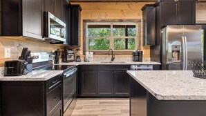 A modern kitchen with dark cabinetry, stainless steel appliances, granite countertops, and a window providing a view of greenery.