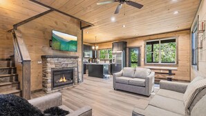 A spacious living area with a fireplace, a ceiling fan, and a kitchen in the background.