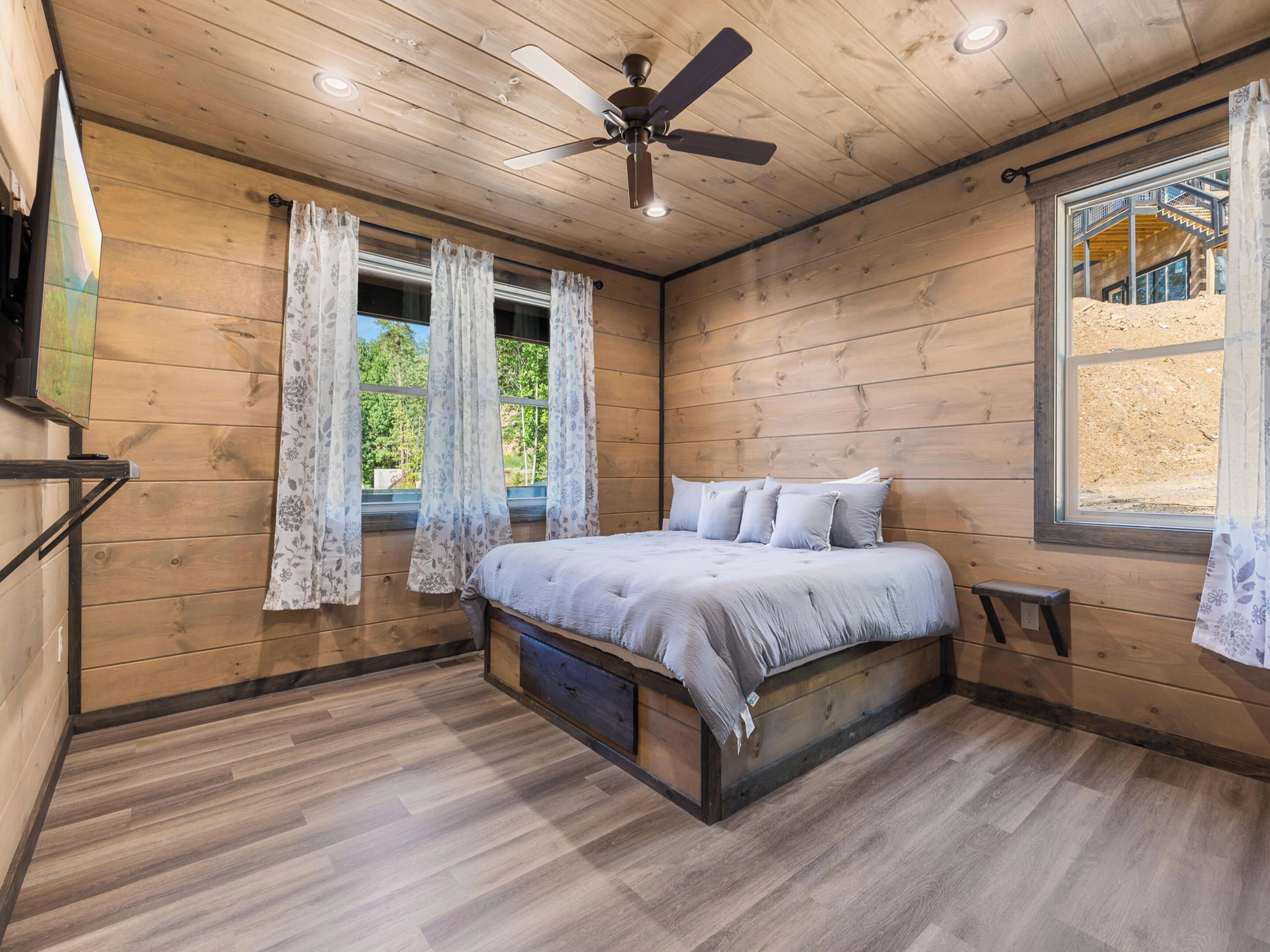 A wooden cabin bedroom with a bed, curtains, a ceiling fan, and a window.