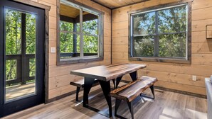 A wooden cabin interior with a large table and bench, large windows, and a view of trees.