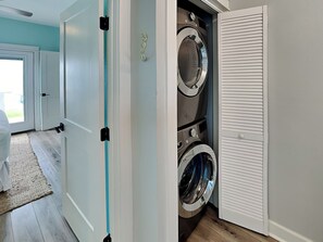 A laundry room with two stacked front-loading washing machines, a door with a handle, and a view into a room with a door and a window. - Copano Coastal Gem by Miss Kitty’s (Rockport)